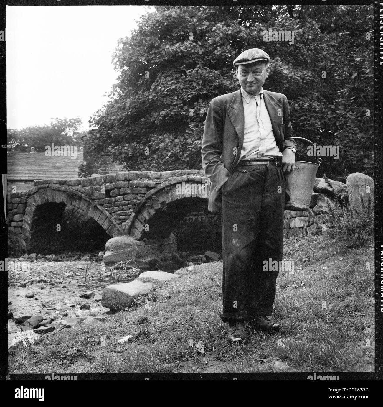 Ein informelles Porträt eines Mannes, der traditionelle Clogs trägt, einen Eimer hält, der vor der Packhorse Bridge in Wycoller, Trawden Forest, Pendle, Lancashire, Großbritannien, steht. Stockfoto