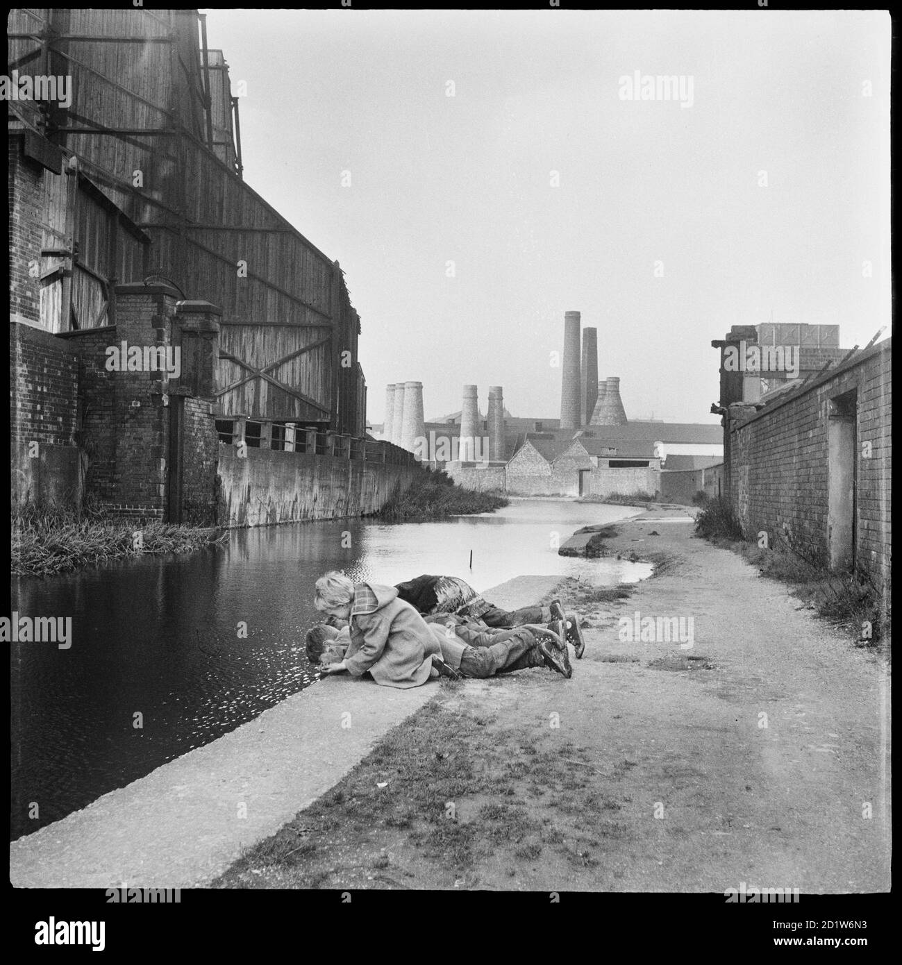 Kinder liegen auf dem Treidelpfad mit Blick in das Wasser des Caldon-Kanals gegenüber der Elektrizitätswerke mit Trent Works und Westwood Mills im Hintergrund, Caldon Canal, Joiner's Square, Hanley, Stoke-on-Trent, Staffordshire, Großbritannien. Stockfoto