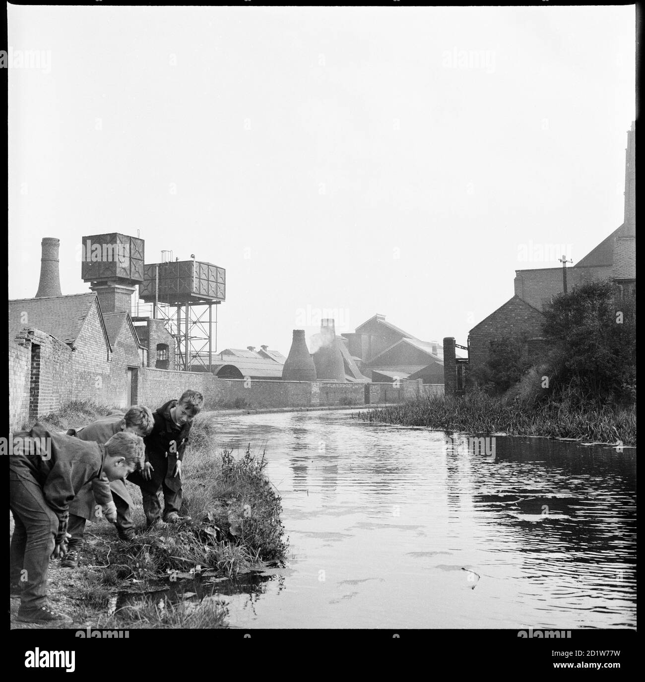 Drei Jungen, die das Wasser des Caldon-Kanals von einem Punkt auf dem Schlepppfad neben Trent Works, mit den Öfen von Joiners Square Mill im Hintergrund, Joiner's Square, Hanley, Stoke-on-Trent, Staffordshire, Großbritannien. Stockfoto