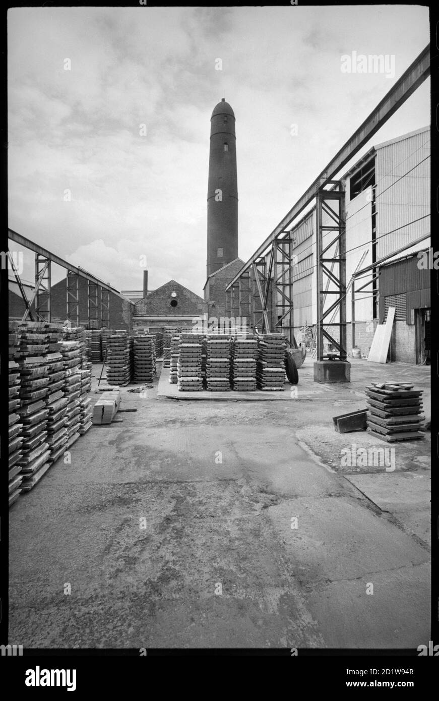 Allgemeine Ansicht des Elswick Shot Tower, abgerissen 1968-69, verwendet für die Erstellung von Bleiaufnahmen, mit einem Hof mit Paletten im Vordergrund Schusstürme wurden verwendet, um sphärische Aufnahmen von Blei zu erstellen. Dies wurde getan, indem man geschmolzenes Blei durch einen Seicve hinunter einen großen Schacht oder Turm in Wasser fallen ließ. Das Metall kühlt ab, wenn es fällt und dreht sich aufgrund der Oberflächenspannung kugelförmig. Dieser Schussturm war Teil der Elswick Bleiwerke, und der Turm wurde abgerissen 1968-69. , um 1955 - um 1969. Stockfoto