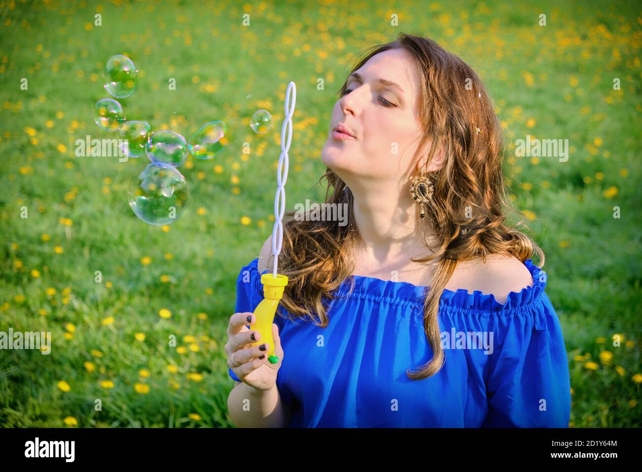 Junge Frau in einem blauen Kleid bläst Seifenblasen auf Ein Hintergrund eines Feldes mit Löwenzahn Stockfoto