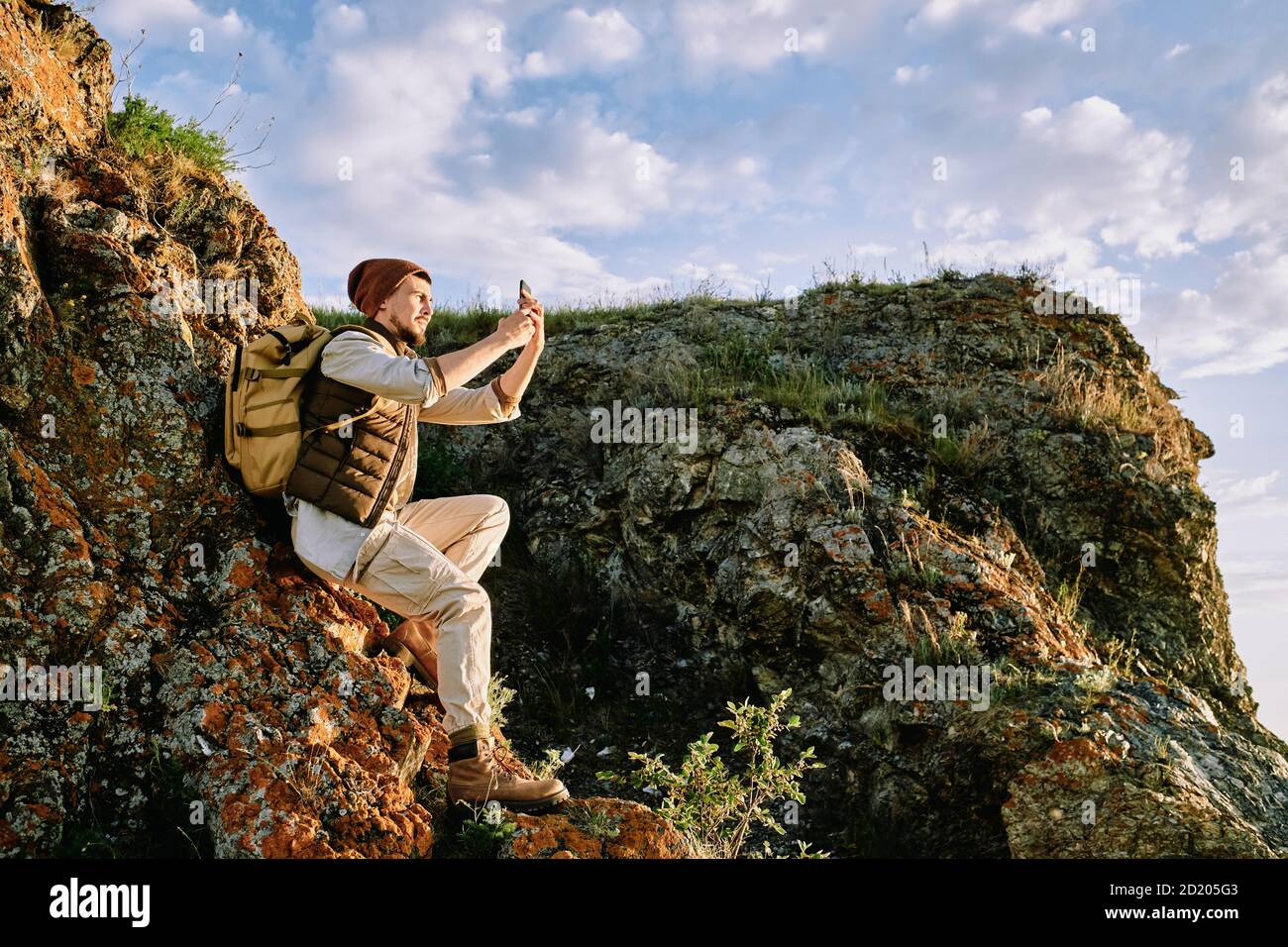 Schöner bärtiger Wanderer in Weste sitzt auf Felsen und Felsen Landschaftsfotografie auf dem Smartphone Stockfoto