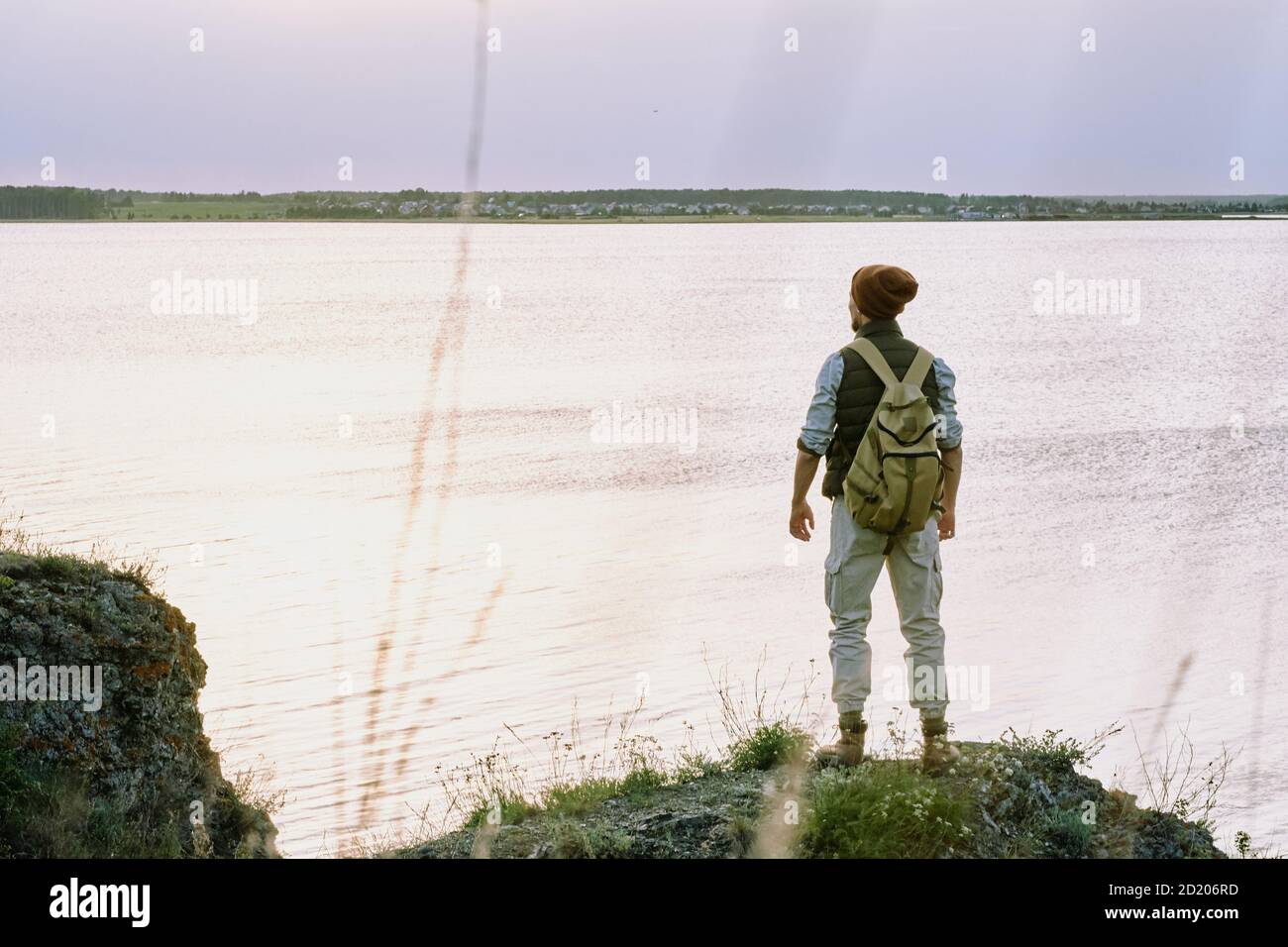 Rückansicht des jungen männlichen Rucksacktouristen im Hut stehend auf Am Rand der Klippe und dem See während der Wanderung genießen Stockfoto