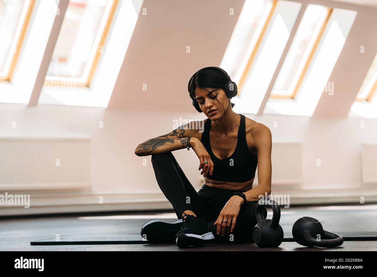 Junge schlanke Frau mit Kopfhörern, die nach dem Training eine Pause einlegen Mit Kettlebells im Fitnessstudio Stockfoto