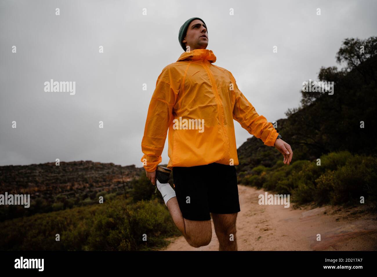 Hübscher junger Trailrunner, der die Beinmuskulatur vor dem Laufen dehnt Bergweg bei bewölktem Wetter mit Büschen Stockfoto