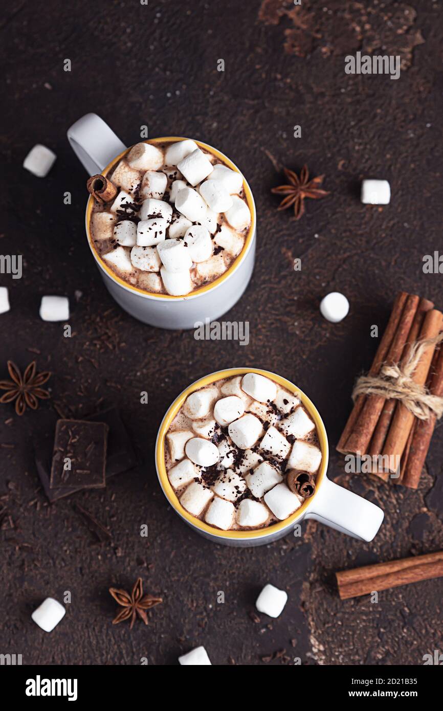 Zwei weiße Keramikbecher mit heißem Kakao oder Schokolade mit Marshmallow und Anis, Zimt und Bitterschokolade auf dunkelbraunem Steingrund. Stockfoto