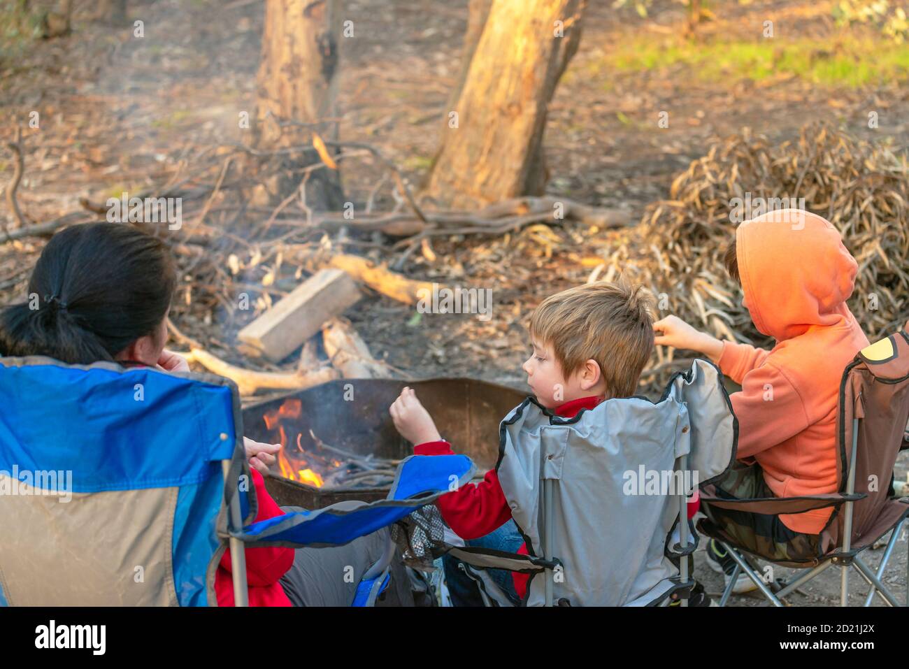 Kinder brennen Feuer auf Kuitpo Wald Campingplatz, während ihre Mutter entspannt in den Schulferien auf dem Stuhl Stockfoto