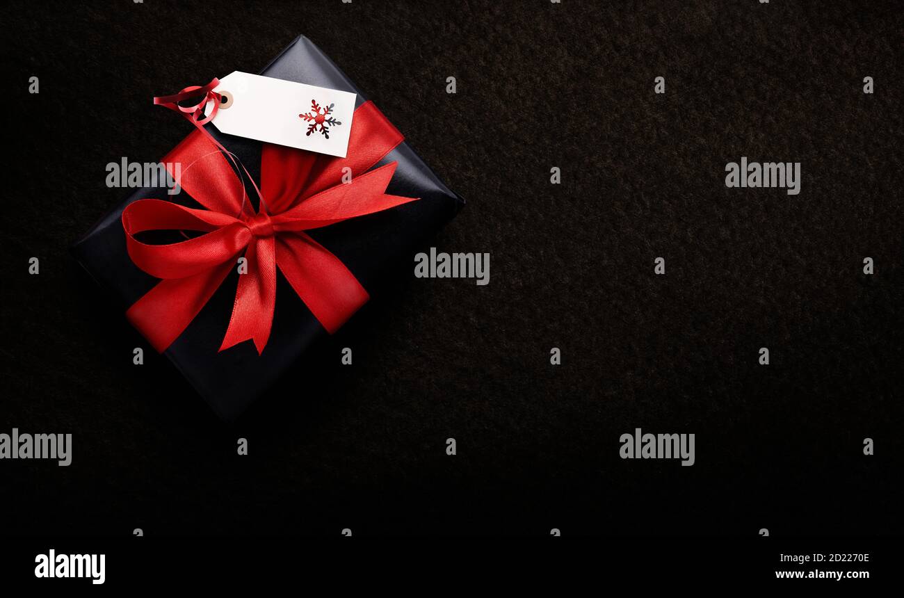 Ein Weihnachtsgeschenk in schwarzem Papier und rotem Band mit einem leeren Tag eingewickelt, Karte vor einem dunklen Hintergrund von oben. Stockfoto
