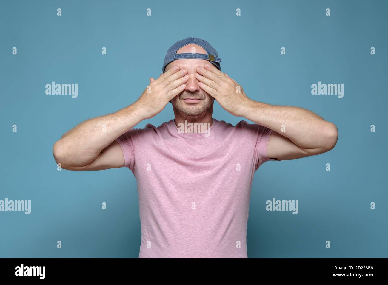 Ruhiger Mann in einer Mütze und ein rosa T-Shirt bedeckte seine Augen mit Händen, er erwartet eine Überraschung oder will nichts sehen. Stockfoto