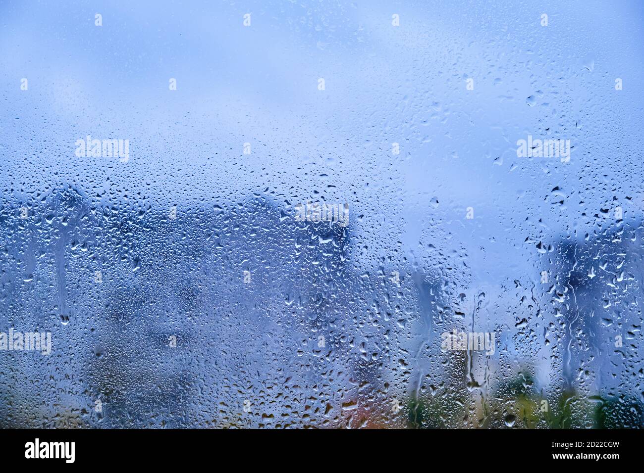 Glas mit Regentropfen auf dem Hintergrund einer verschwommenen Stadt. Herbstfenster mit der Textur des Wassers fällt gegen einen trüben grauen Himmel Stockfoto