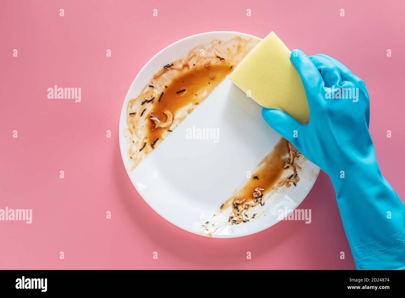 Hand in blauem Gummihandschuh halten gelben Reinigungsschwamm an Reinigen und waschen Sie Lebensmittelflecken und Schmutz auf Weiß Gericht nach dem Essen Mahlzeit isoliert auf rosa Stockfoto