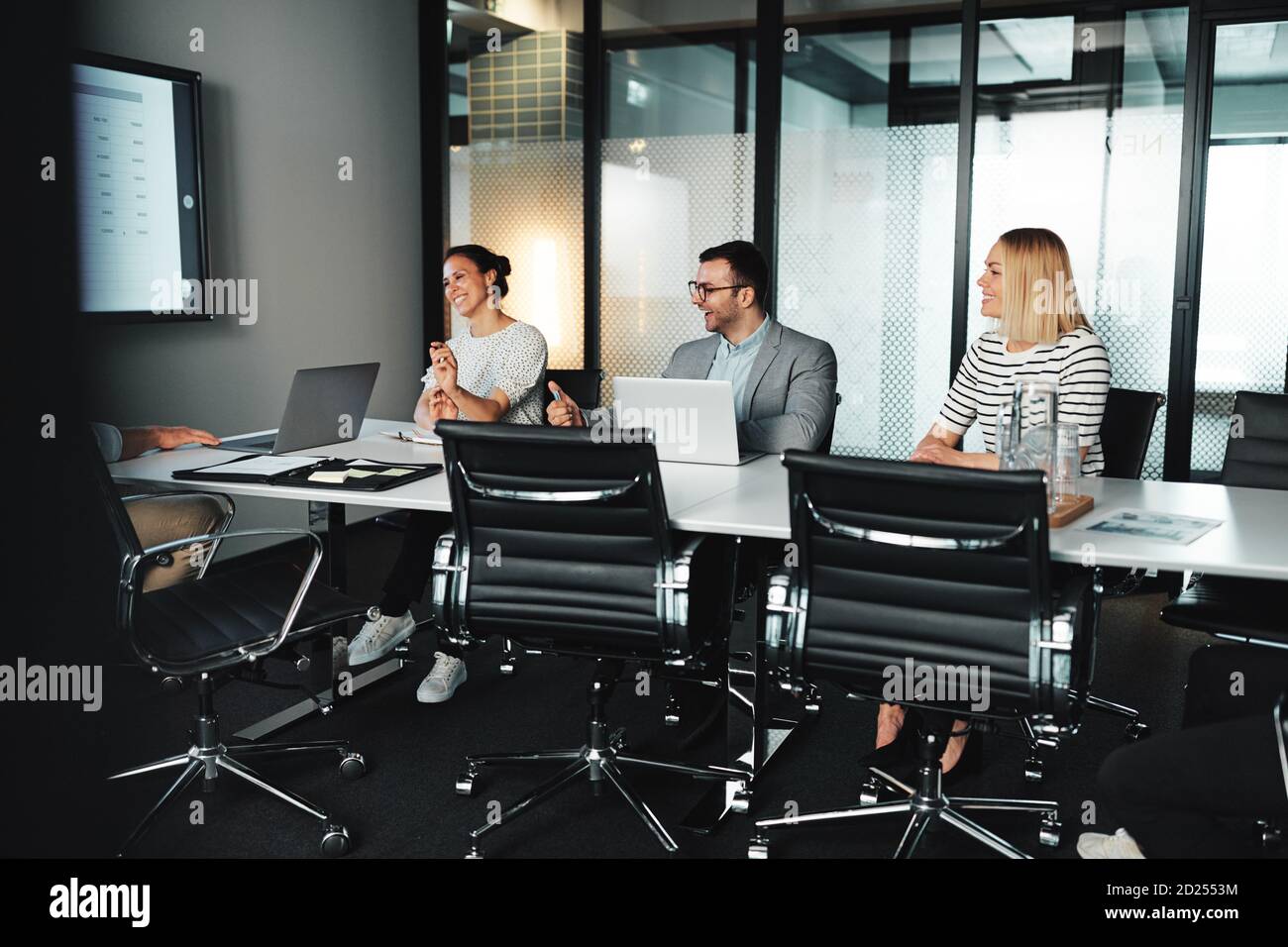 Lächelnde Geschäftsleute, die einer digitalen Präsentation lauschen, während sie herumsitzen Ein Tisch während einer Sitzung im Sitzungssaal Stockfoto