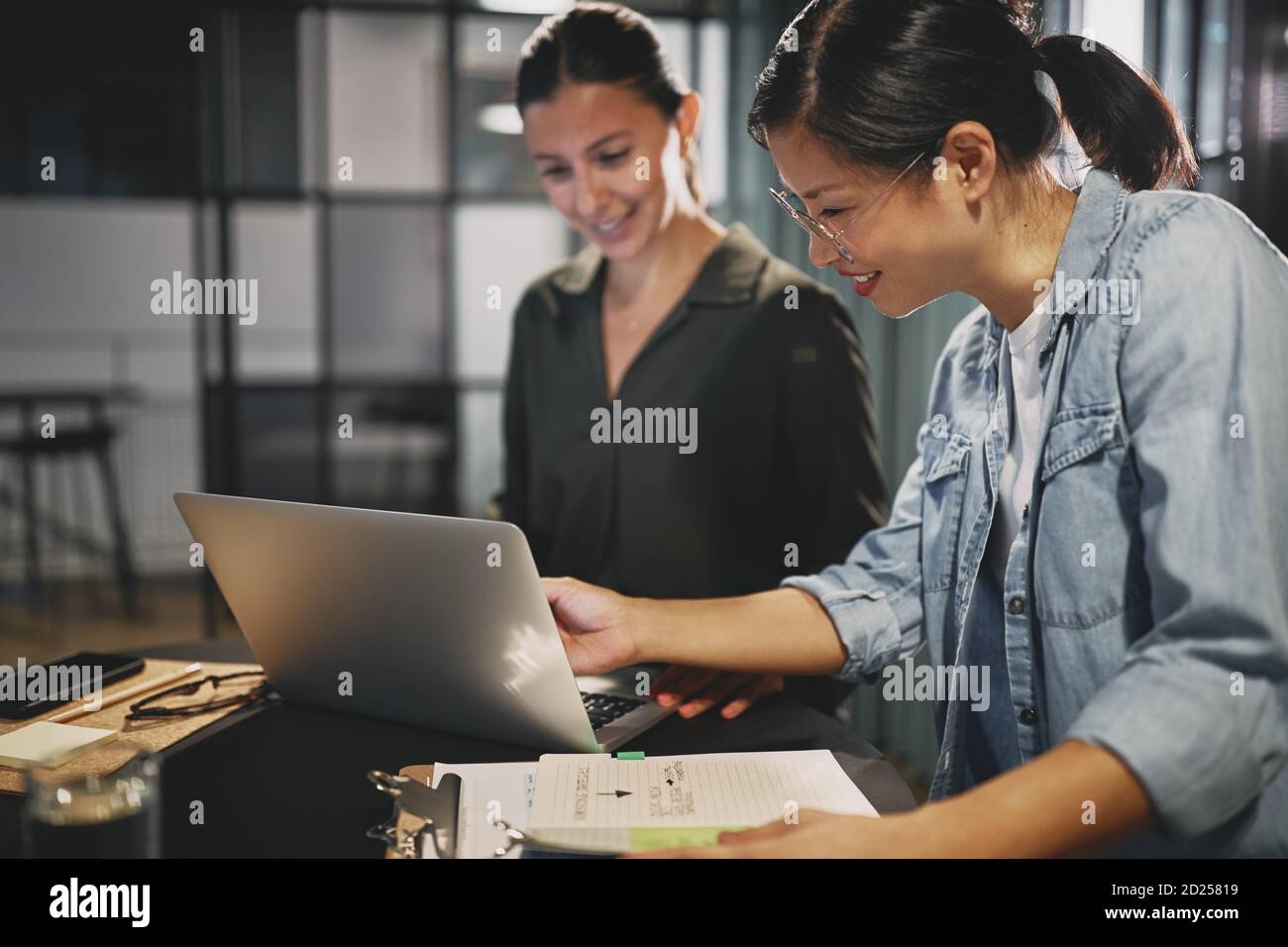 Zwei junge Geschäftsfrauen, die lächeln, während sie an einem Laptop arbeiten Gemeinsam an einem Tisch in einem modernen Büro Stockfoto