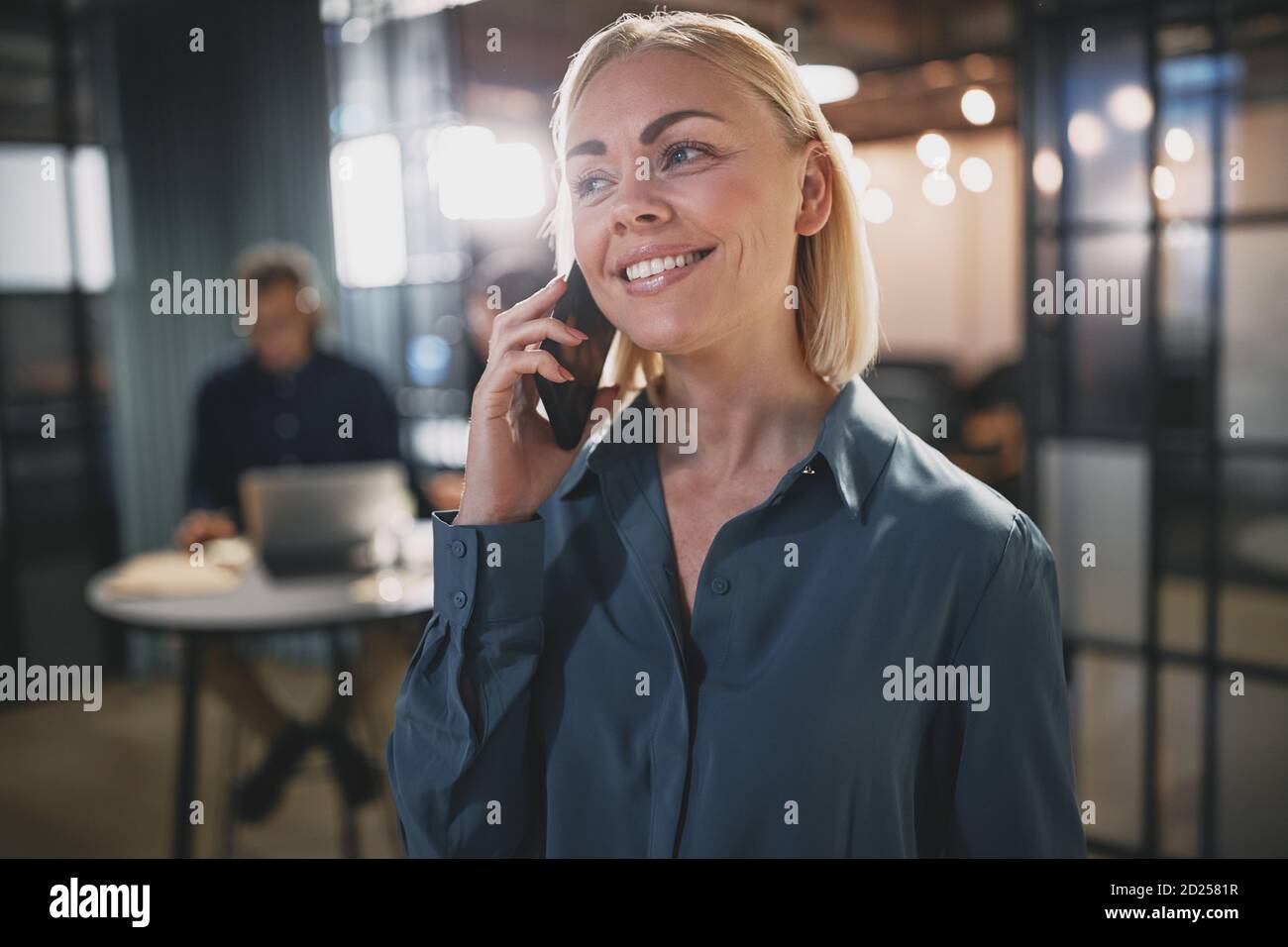 Junge Geschäftsfrau lächelt und spricht auf ihrem Handy, während sie steht In einem Büro mit Kollegen im Hintergrund Stockfoto