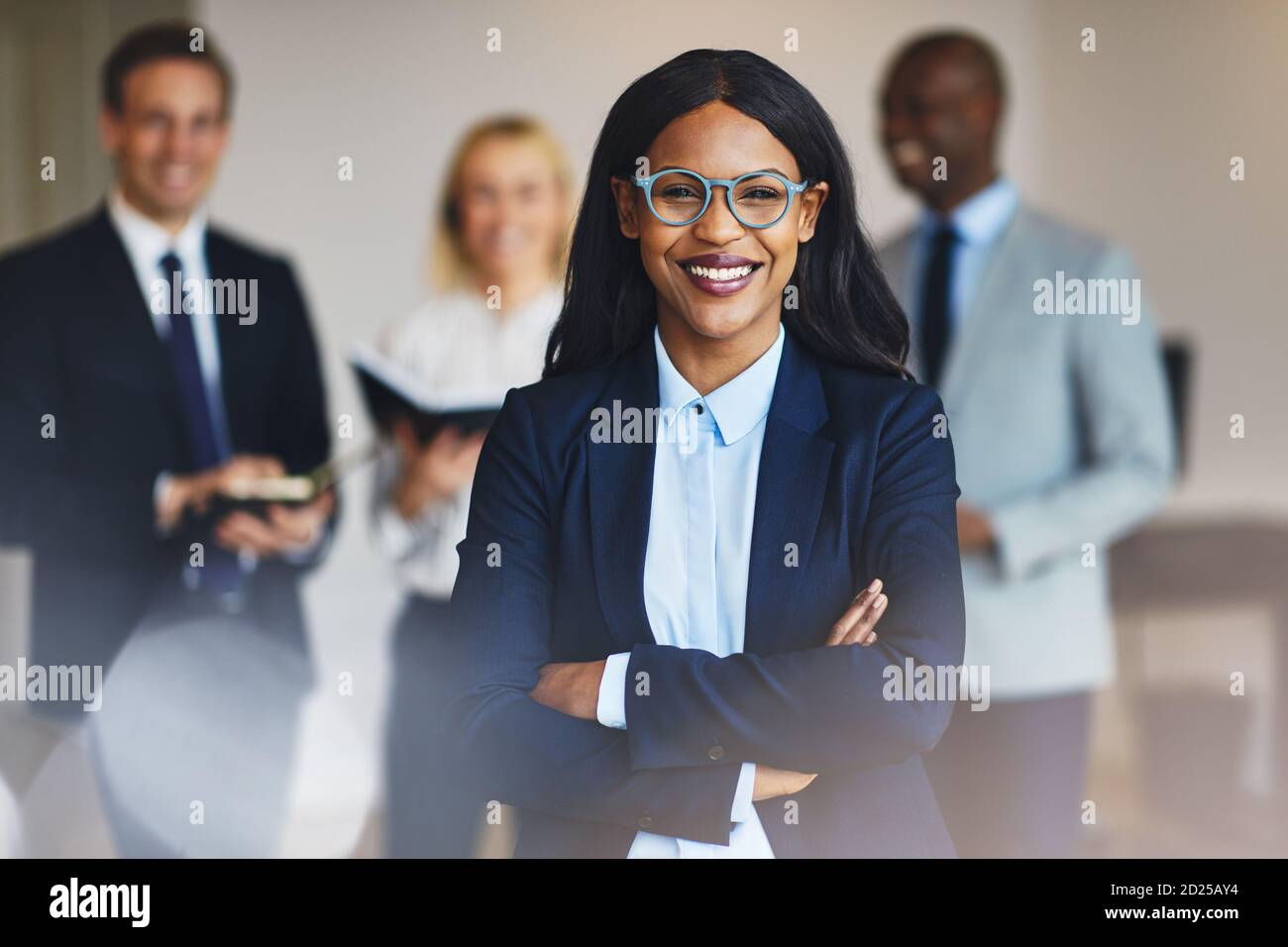 Junge afrikanische Geschäftsfrau stehend mit gekreuzten Armen in einem Büro und lächeln mit Kollegen im Hintergrund stehen Stockfoto