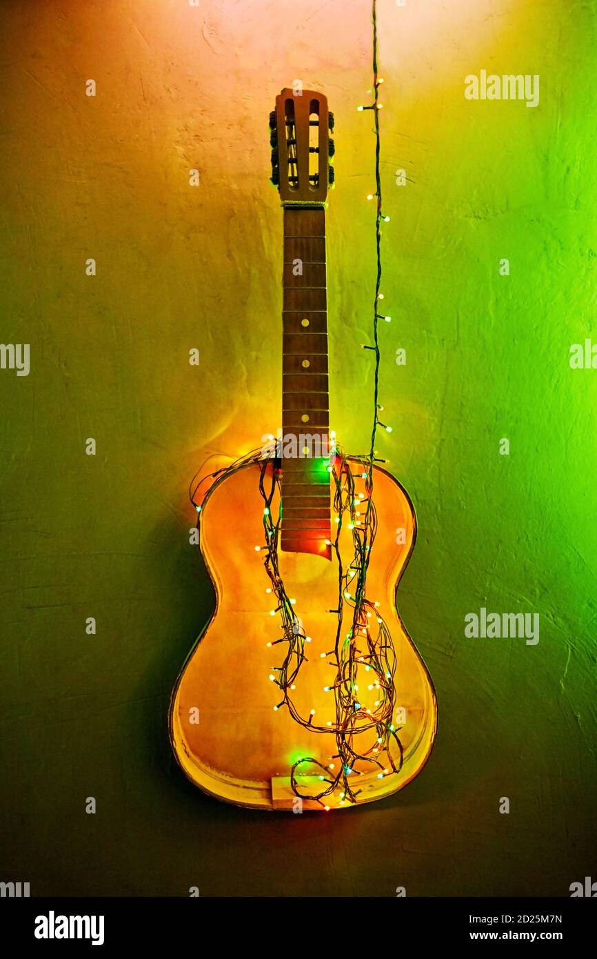 Alte gebrochene Holzgitarre dekoriert leuchtend grün leuchtende Girlande. Musikinstrument, das an der Wand hängt. Weihnachtliche Innenausstattung. Gelb New Yea Stockfoto