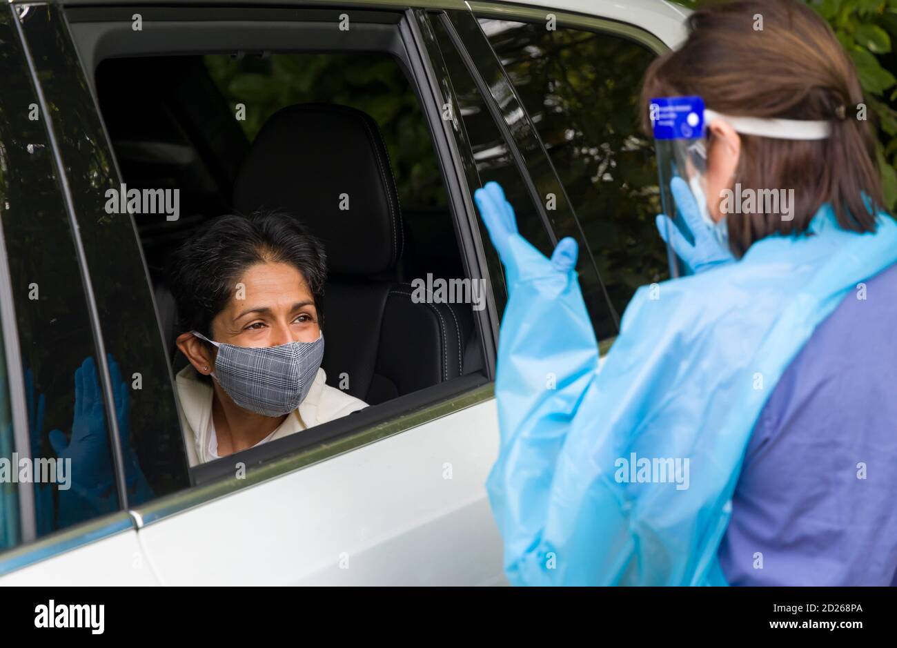 Coronavirus- oder COVID-19-Prävention in einem Testzentrum in England, Großbritannien. Asiatische Frau in einem Auto trägt eine Gesichtsmaske, im Gespräch mit einer Krankenschwester in voller PSA-Ausrüstung. Stockfoto