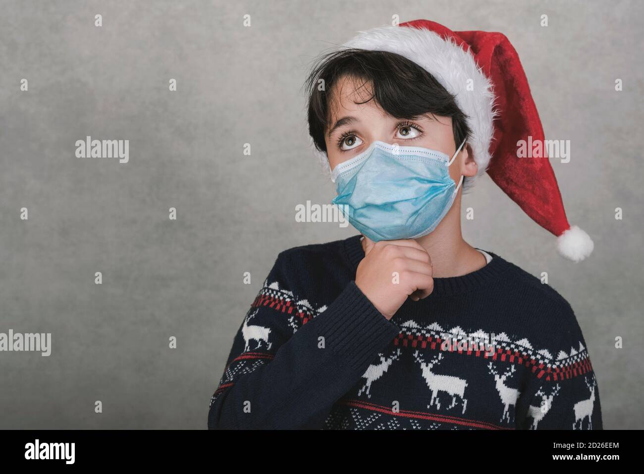 Frohe Weihnachten, nachdenkliches Kind mit medizinischer Maske trägt Weihnachtsmann Hut Auf grauem Hintergrund Stockfoto