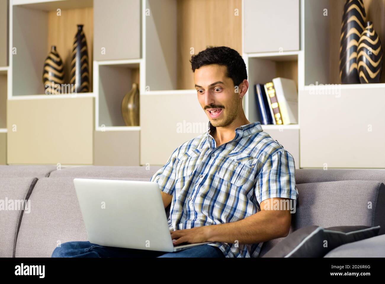 Junger Mann reagiert erstaunt auf einen Laptop-Computer starren Mit breitem Auge mit seinem Mund agape, als er sich zu Hause entspannt Auf einem bequemen Sofa Stockfoto