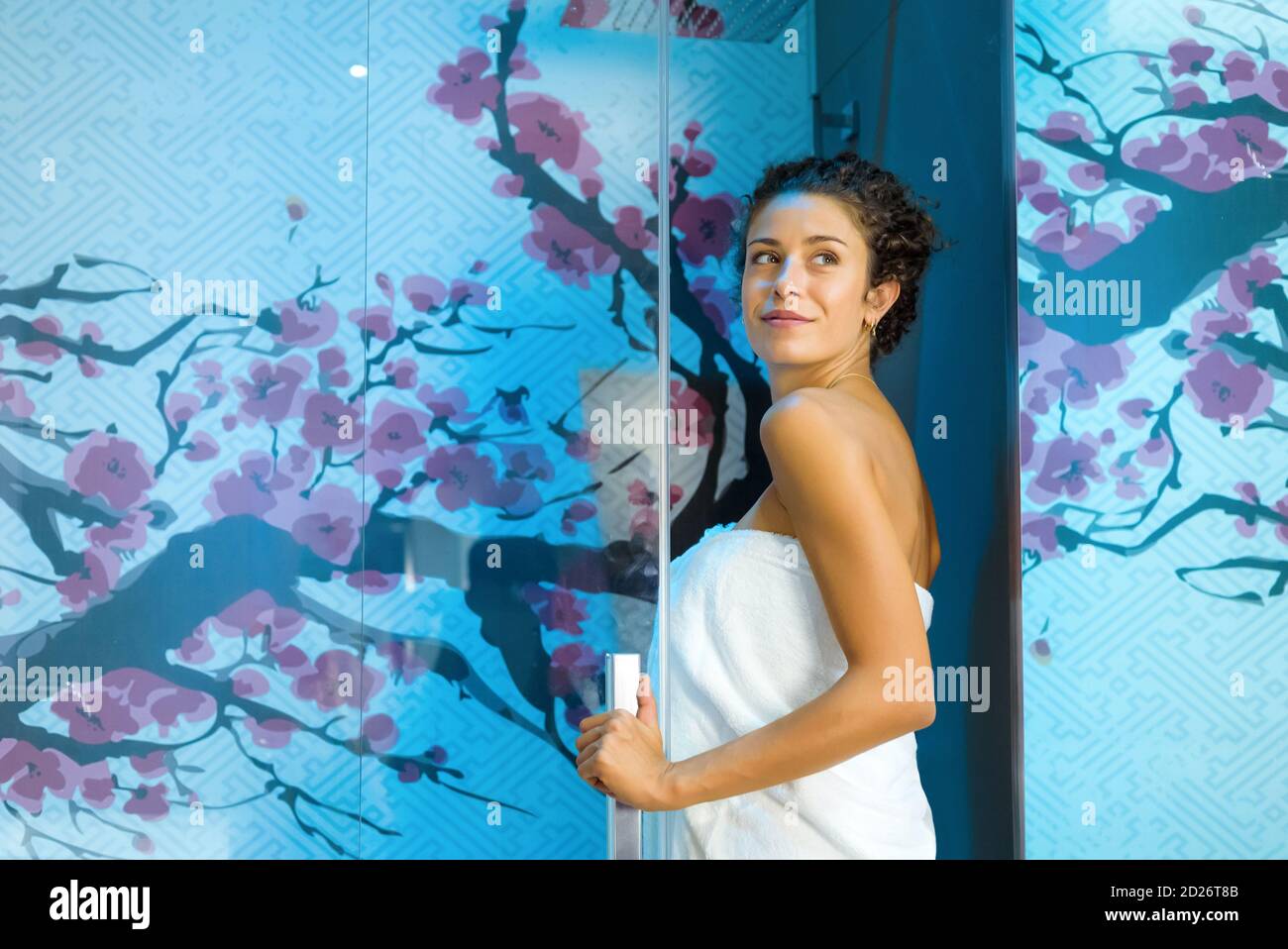Junge Frau betritt eine farbenfrohe blaue Duschkabine und schaut nach hinten Sie, als sie durch die Glastür in einem tritt Konzept der persönlichen Hygiene Stockfoto