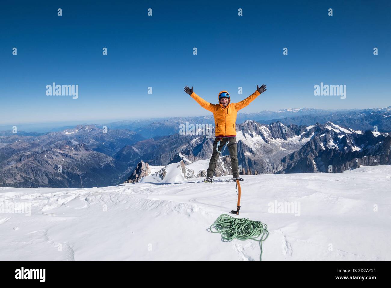 Glücklich lächelnder Seilmannschaftmann mit Kletteraxt gekleidet orange Bergsport Kleidung, Stiefel mit Steigeisen und Sicherheitsgeschirr eine Hände aufgesetzt Auf dem Mont B Stockfoto