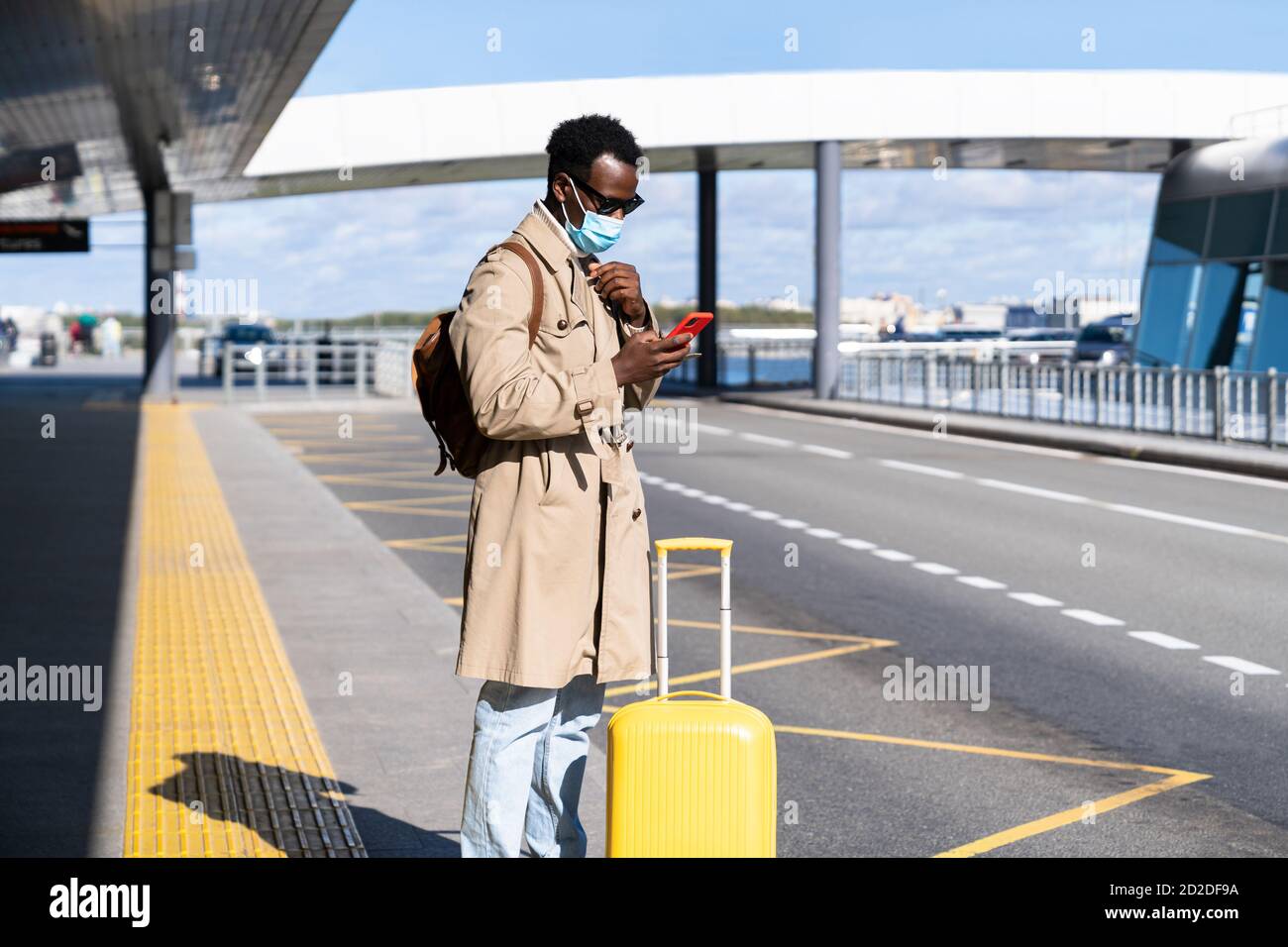 Afro-amerikanische Millennial Traveler Mann mit Koffer steht im Flughafen-Terminal, hält Handy, rufen für ein Taxi oder Car Sharing. Schwarzer Stecker in Stockfoto