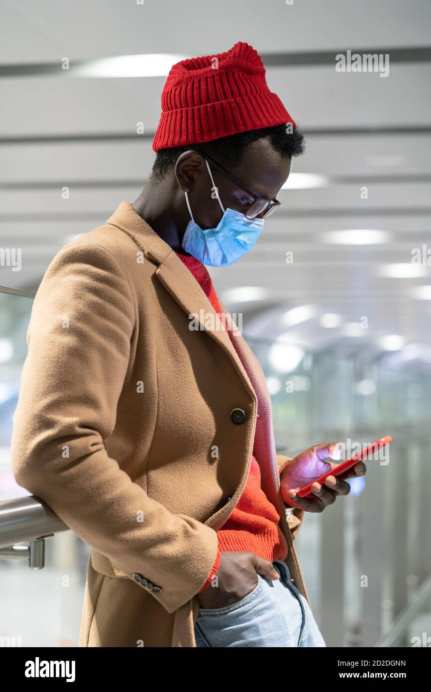 Stilvolle schwarze Reisende Millennial Mann tragen Gesicht Schutzmaske, stehend in der Halle des Flughafens, mit Handy. Covid-19, Coronavirus der zweiten Welle, n Stockfoto