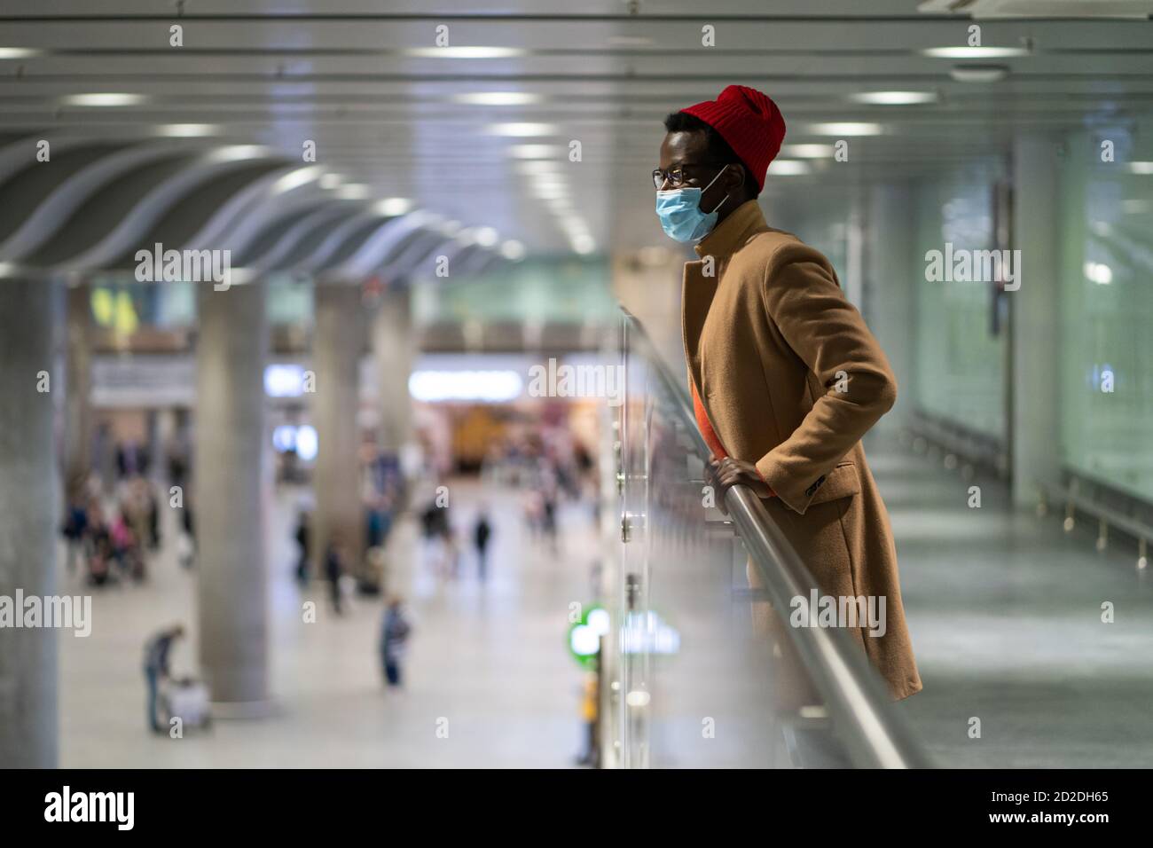 Seitenansicht der stilvollen Black Traveler Millennial Mann tragen Gesicht Schutzmaske, stehend in der Halle des Flughafens, Blick nach unten, verschwommener Hintergrund. Covid-19, Stockfoto