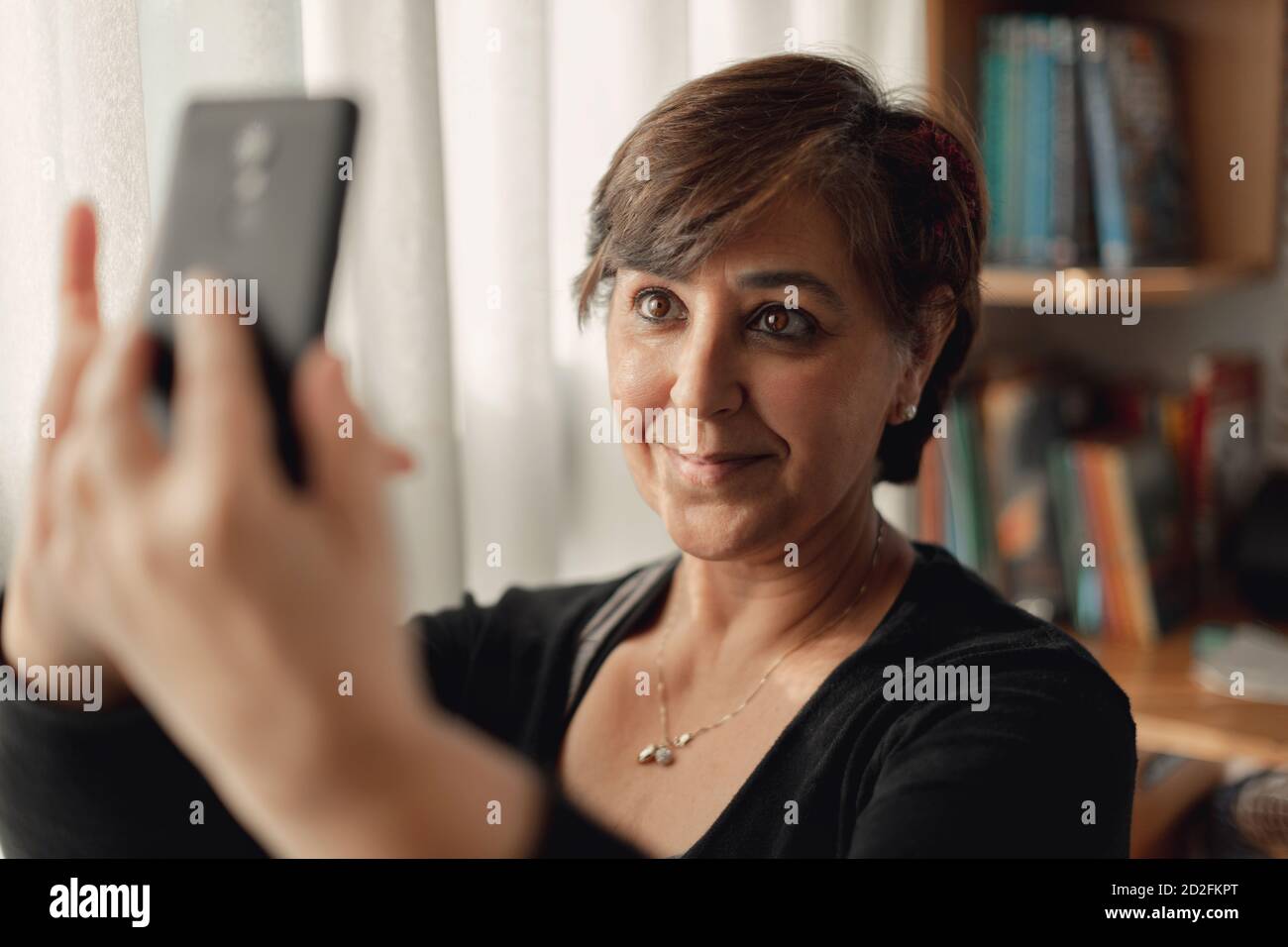 Erwachsene Frau mit kurzen Haaren macht Selfies mit ihrem Smartphone in ihrem Studio. Zu Hause in Quarantäne bleiben und Zeit im Sonnenlicht verbringen. Stockfoto