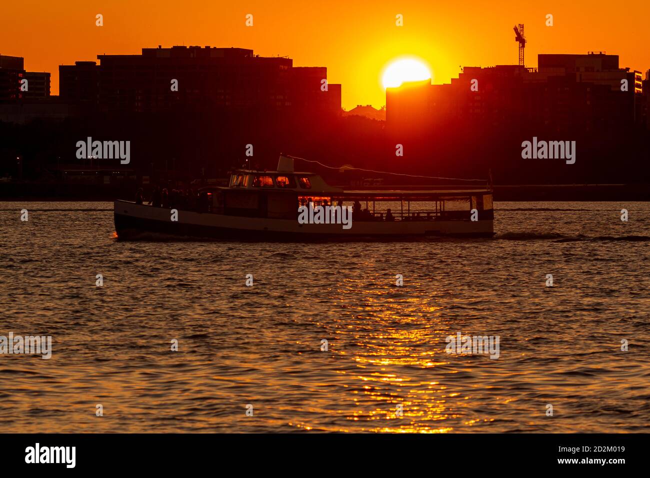 Ein Sonnenuntergangsbild mit Sonnenuntergang hinter Gebäuden, während ein Flusskreuzfahrtschiff mit Passagieren an Deck im Potomac-Fluss unterwegs ist, der r Stockfoto