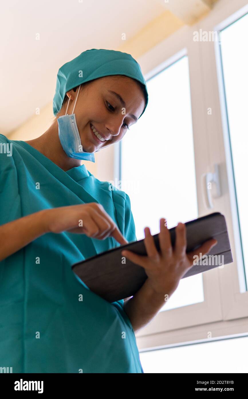 Junge Krankenschwester in blauen Peelings und medizinische Maske Lesen von Informationen Auf Tablette, während im Korridor des modernen Krankenhauses stehend Stockfoto