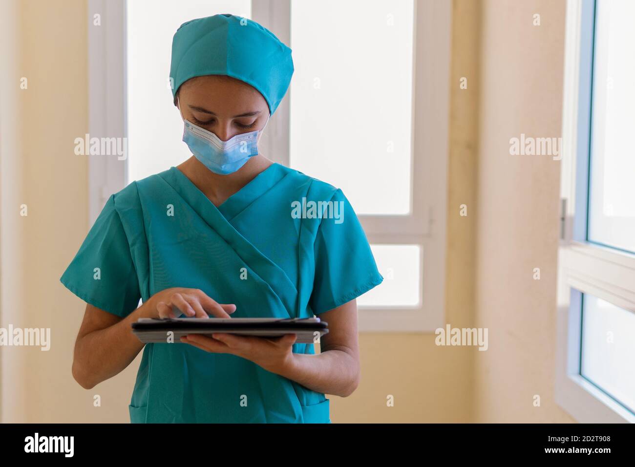 Junge Krankenschwester in blauen Peelings und medizinische Maske Lesen von Informationen Auf Tablette, während im Korridor des modernen Krankenhauses stehend Stockfoto