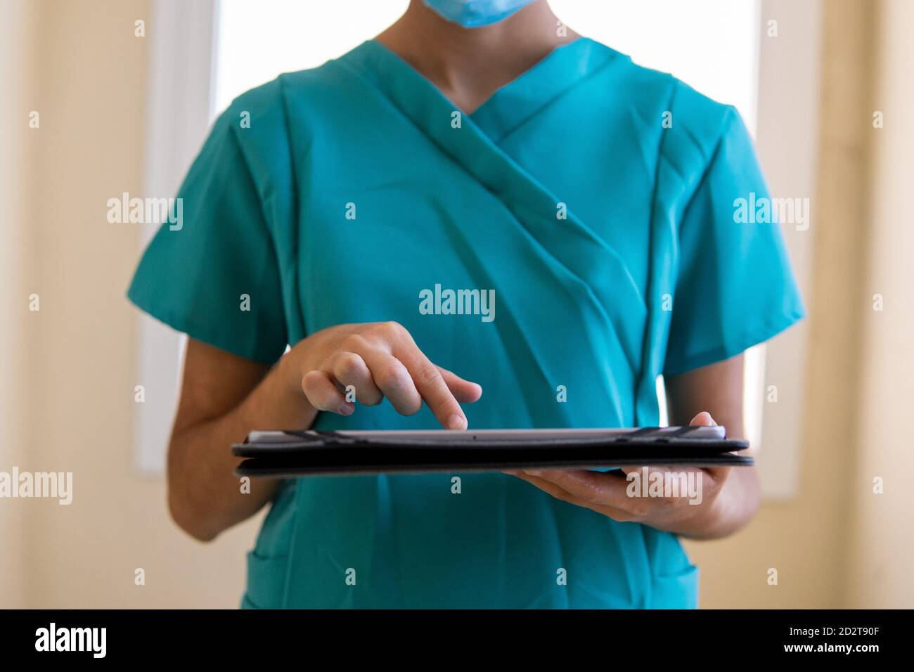 Junge Krankenschwester in blauen Peelings und medizinische Maske Lesen von Informationen Auf Tablette, während im Korridor des modernen Krankenhauses stehend Stockfoto