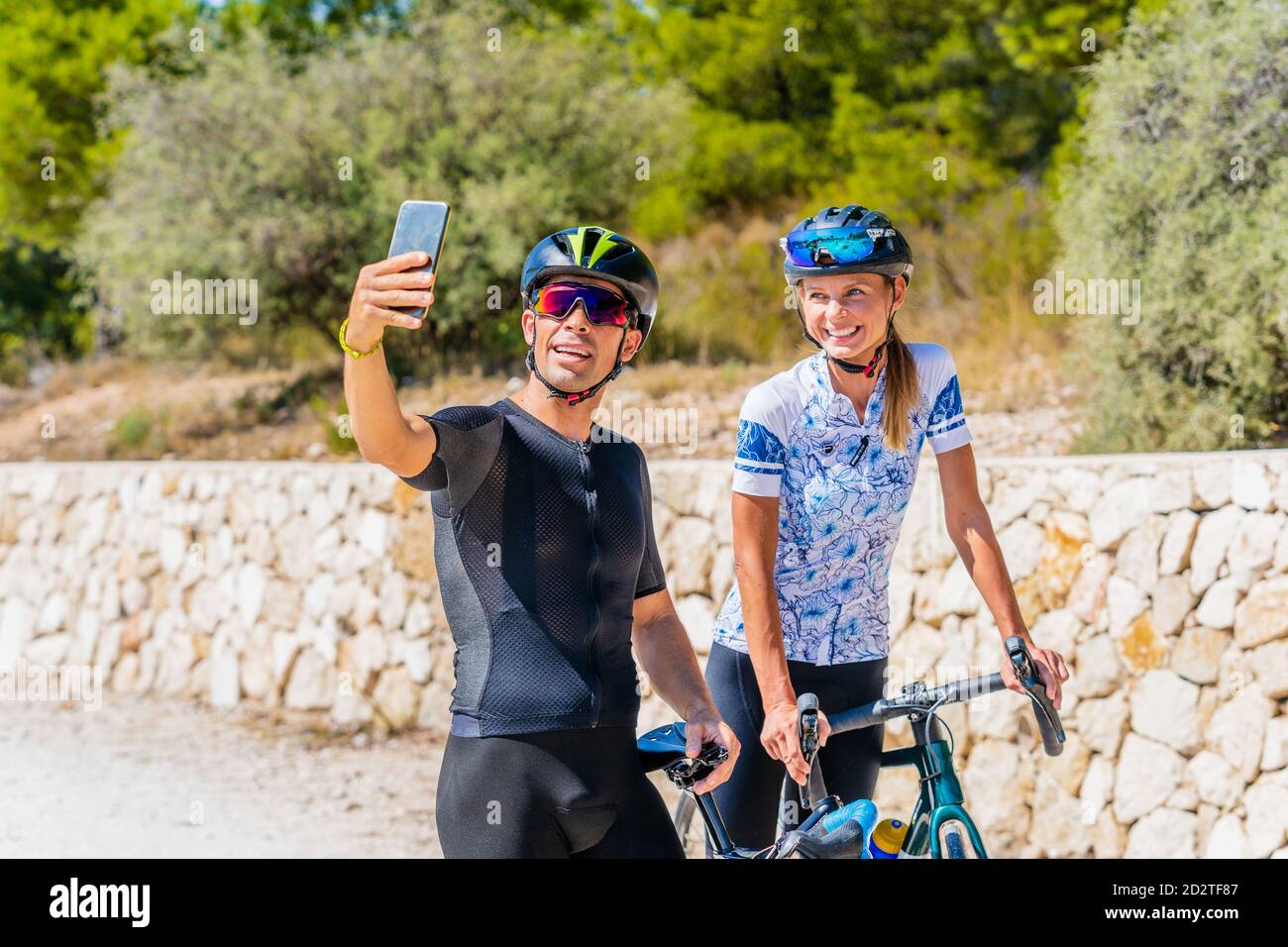 Glückliche junge aktive Mann und Frau in Sportbekleidung und Schutzhelmen Selfie auf dem Smartphone beim Stehen mit Fahrrädern auf der Landstraße und Vorbereitung auf das Reiten Stockfoto
