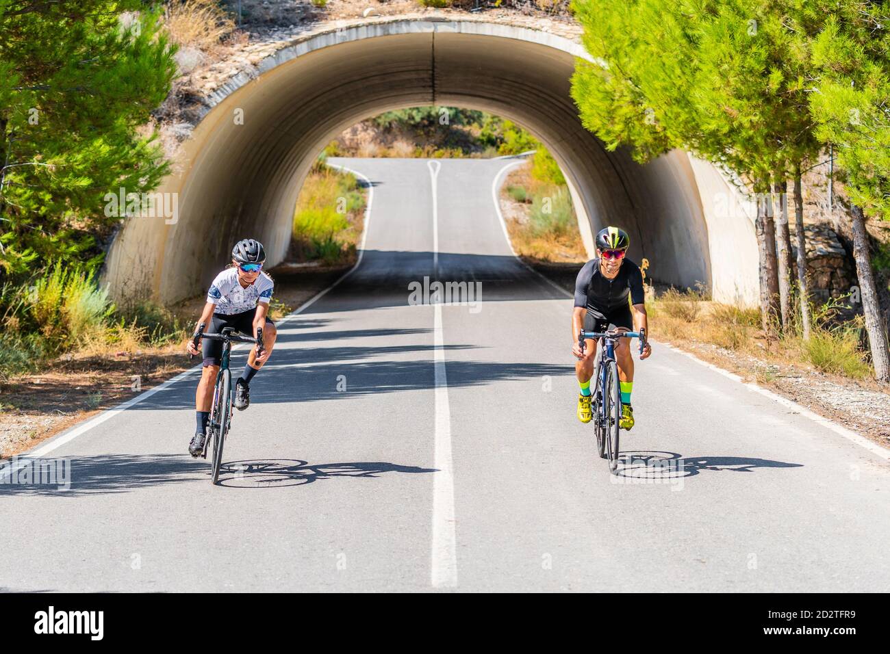 Die ganze Länge der Radfahrer in Sportbekleidung und Helme zusammen Radfahren Auf asphaltierter Fahrbahn unter gewölbter Brücke in der Sommerlandschaft Stockfoto
