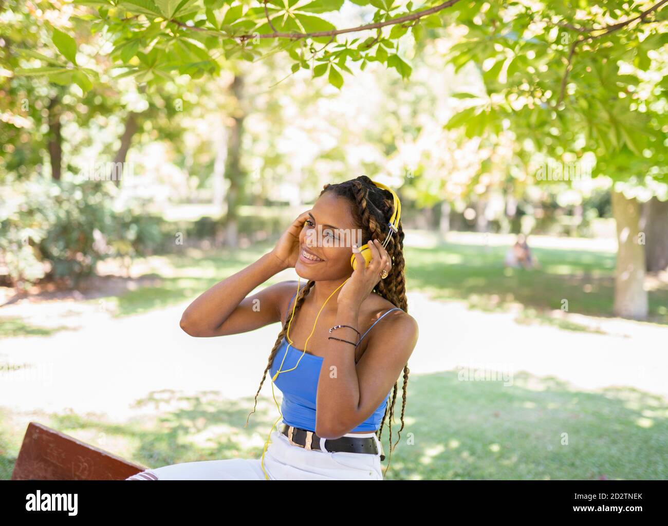 Positive hispanische Frau, die im Stehen Musik in Kopfhörern hört Im Stadtpark an sonnigen Tag und Blick weg Stockfoto