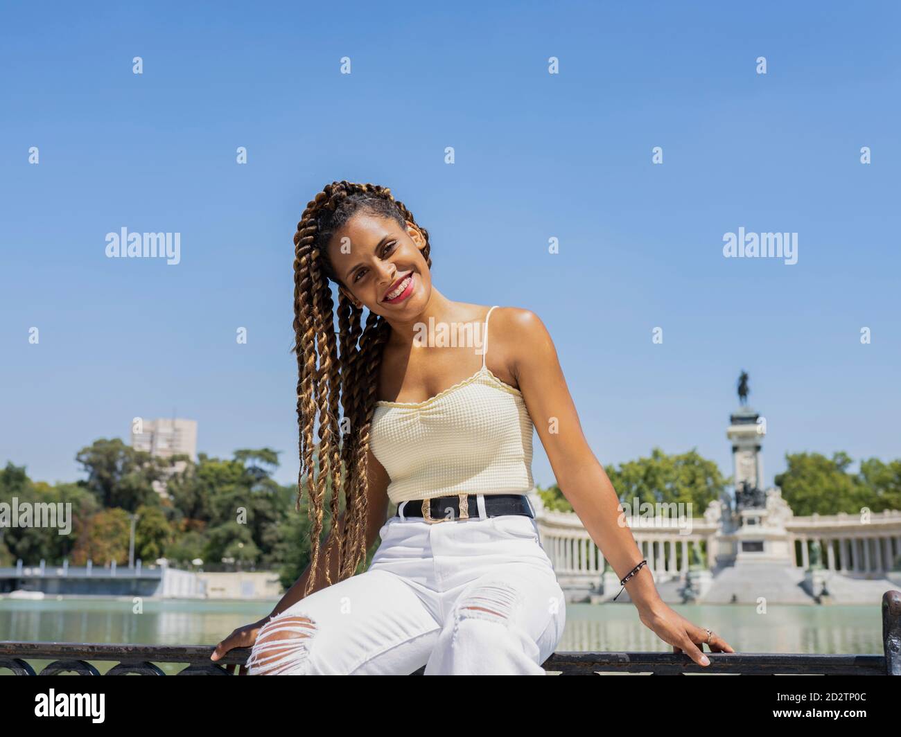Unbeschwerte hispanische Hündin im trendigen Outfit und mit Zöpfen sitzend Zaun in der Nähe von See in Stadtpark und Blick auf Kamera Stockfoto
