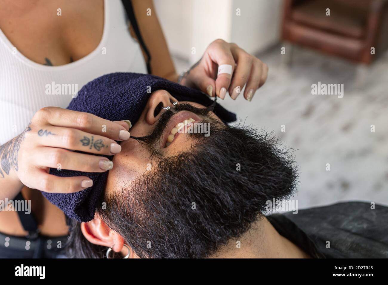 Hoher Winkel der Ernte unkenntlich weibliche Master mit tätowierten Körper Styling Schnurrbart von glücklich brutal bärtigen Mann mit Piercing während Haarschnitt Verfahren im Friseurladen Stockfoto