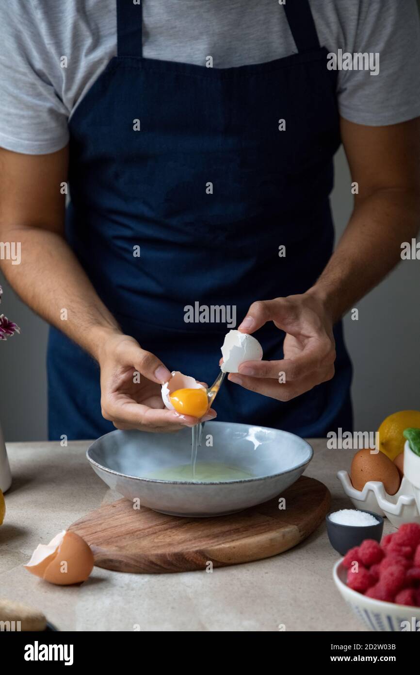 Ernte unkenntlich Mann in Schürze zerbrechen rohes Ei in Schüssel Beim Zubereiten des Frühstücks am Tisch mit Zutaten Stockfoto