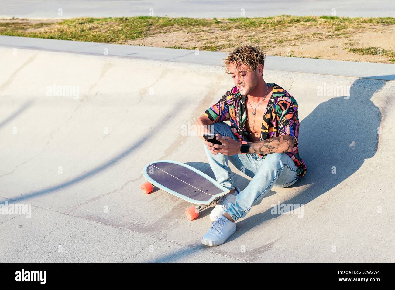 Handsome Millennial lockig behaart Kerl in unknopfed bunte Hemd und Jeans  sitzen mit Skateboard auf Betonrampe und Surfen mobil Telefon während der  Ruhe im Skatepark im Sommer Tag Stockfotografie - Alamy