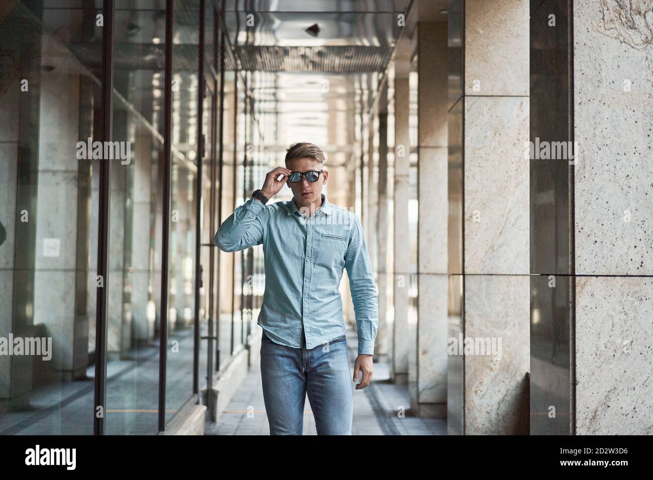 Selbstbewusster Mann in elegantem legerem Outfit, der in der Nähe des modernen Gebäudes steht In der Stadt und berühren Sonnenbrille Stockfoto