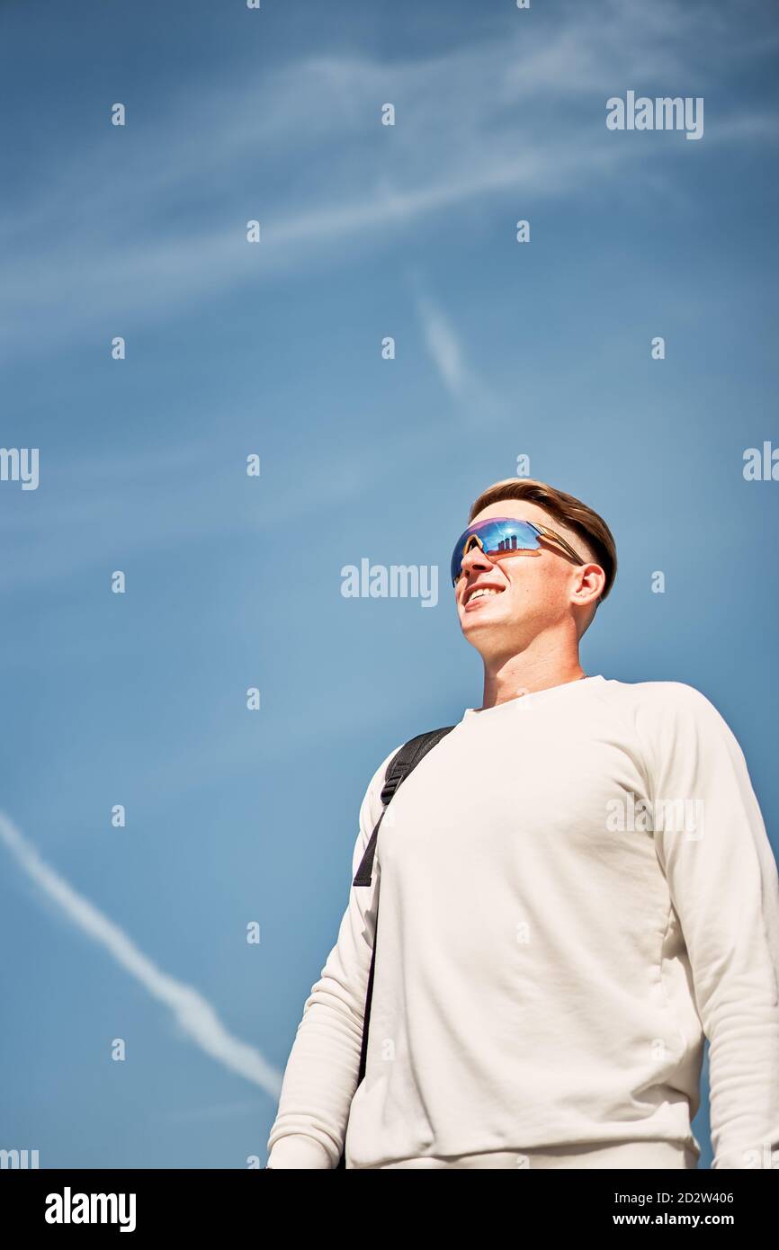 Von unten voller Körper des selbstbewussten jungen männlichen in trendy Lässiges Outfit und Sonnenbrille mit Tasche über der Schulter beim Spazierengehen Städtische Straße mit zeitgenössischen Gebäuden im Hintergrund Stockfoto