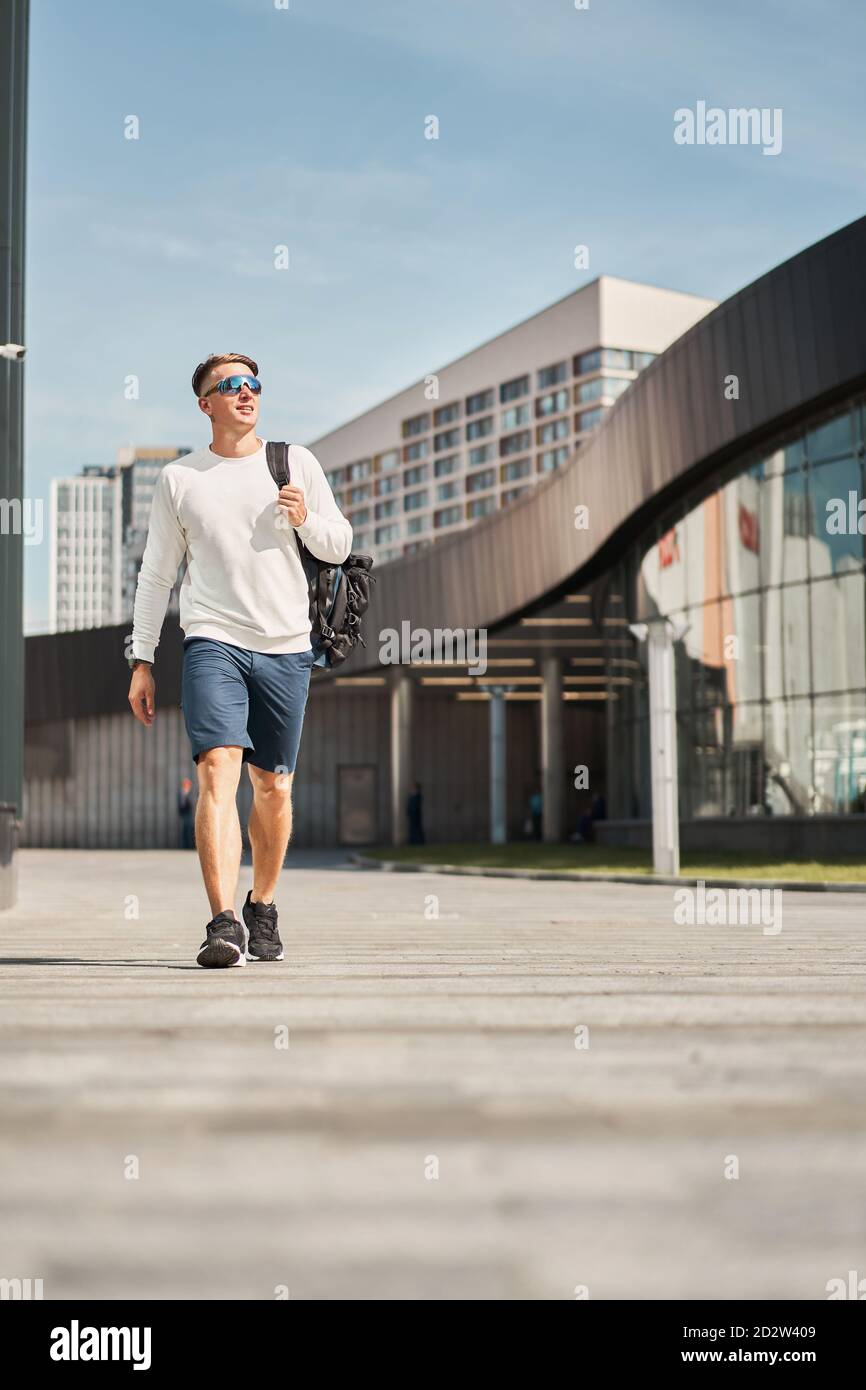 Von unten voller Körper des selbstbewussten jungen männlichen in trendy Lässiges Outfit und Sonnenbrille mit Tasche über der Schulter beim Spazierengehen Städtische Straße mit zeitgenössischen Gebäuden im Hintergrund Stockfoto