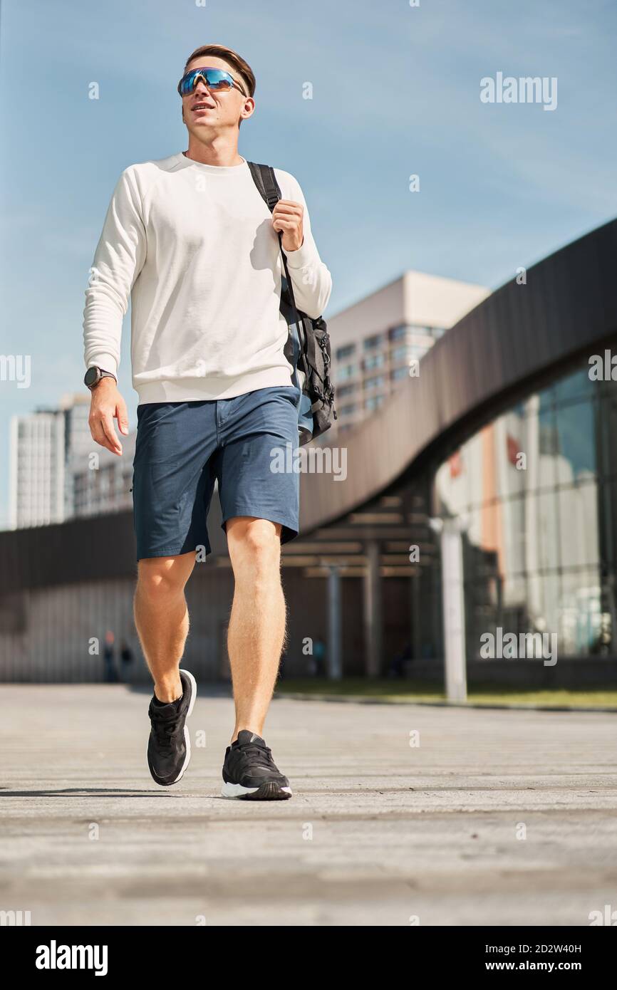 Von unten voller Körper des selbstbewussten jungen männlichen in trendy Lässiges Outfit und Sonnenbrille mit Tasche über der Schulter beim Spazierengehen Städtische Straße mit zeitgenössischen Gebäuden im Hintergrund Stockfoto