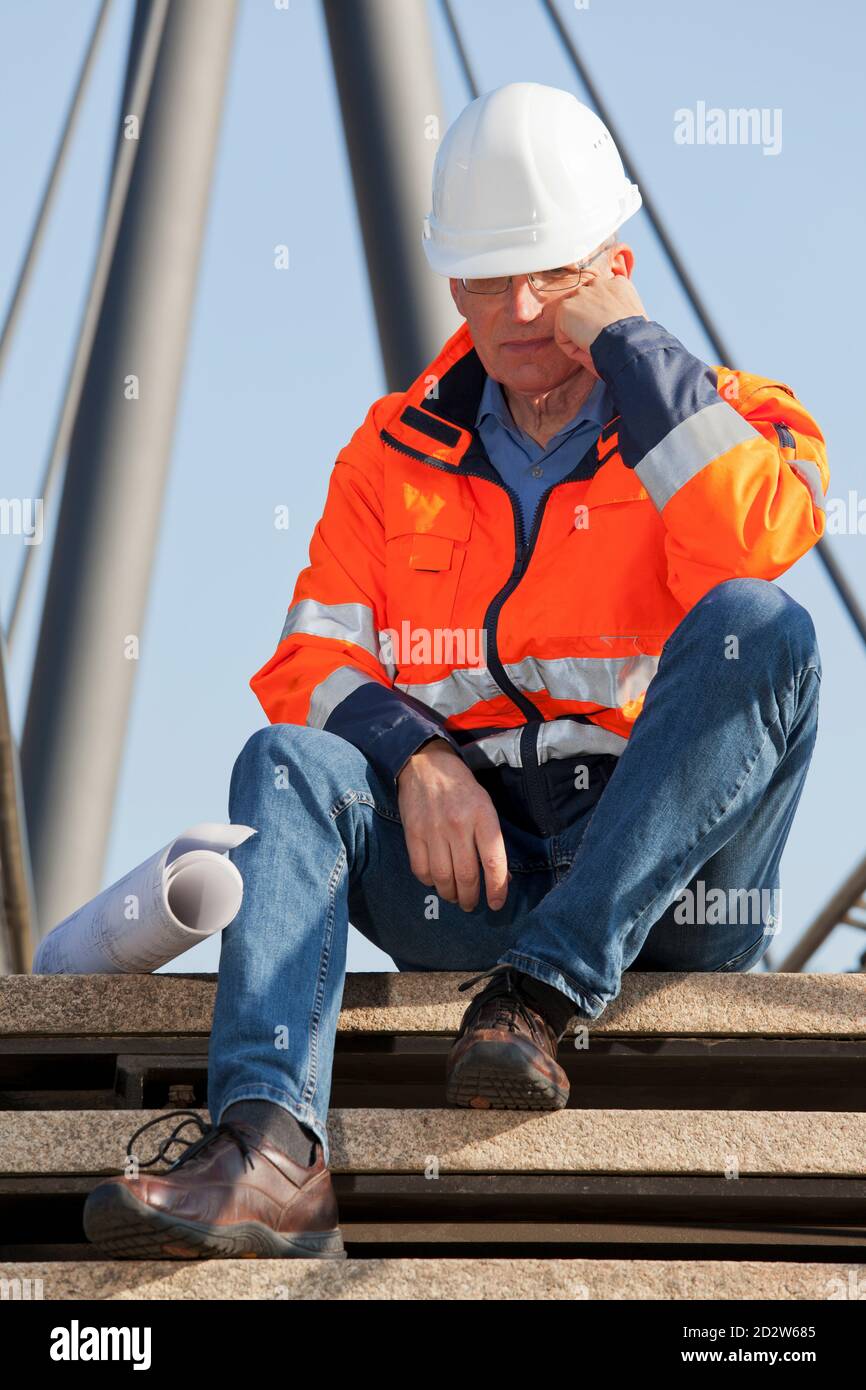 Traurig oder niedergeschlagen Ingenieur oder Vorarbeiter mit Arbeitsschutzkleidung sitzen Vor einem Industriebau - Fokus auf die Gesicht Stockfoto