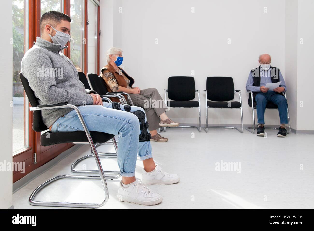 Junge und alte Menschen mit Gesichtsmasken halten soziale Distanz In einem Wartezimmer eines Krankenhauses oder Büros - Fokus auf den jungen Mann im Vordergrund Stockfoto