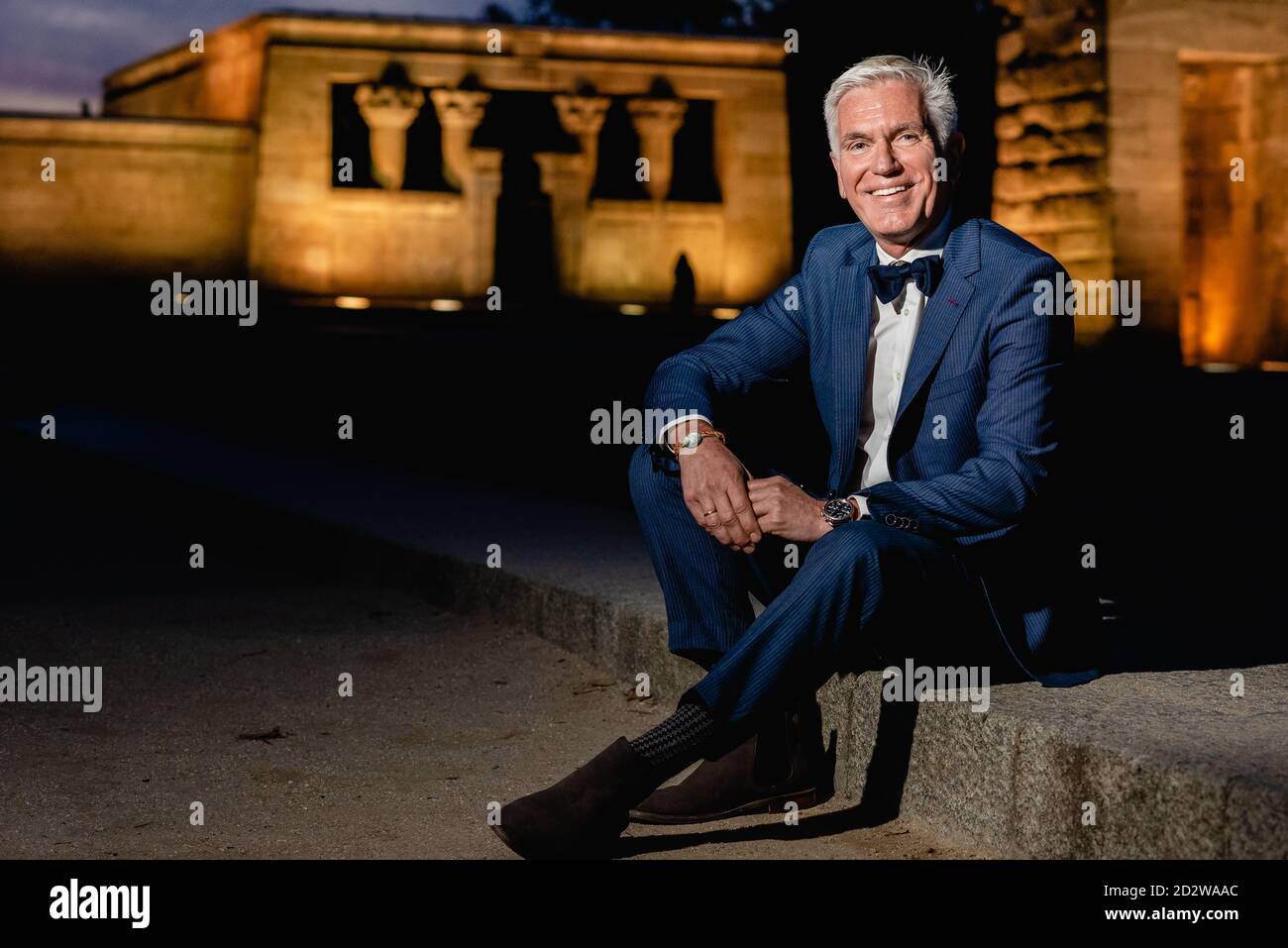 Positiv eleganter Senior-Rüde im stilvollen blauen Anzug mit Schleife Krawatte und Armbanduhren sitzen auf Steinrand in der Stadt der Nacht Und Blick auf die Kamera Stockfoto