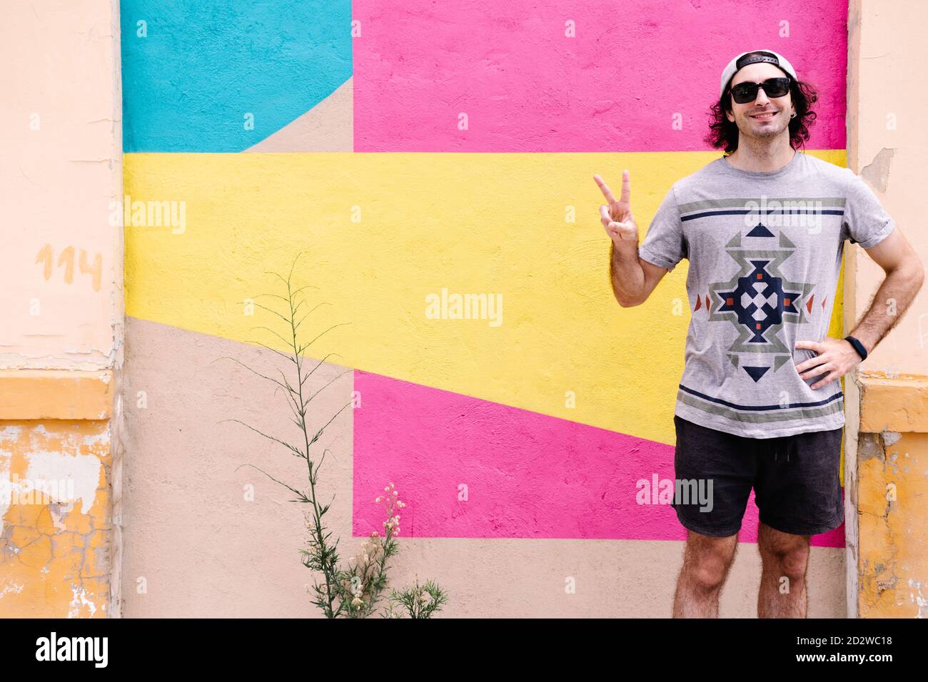 Entzückte junge männliche Hipster in der Nähe von bunten Gebäude in der Stadt Und zeigt V-Zeichen beim Blick auf die Kamera Stockfoto