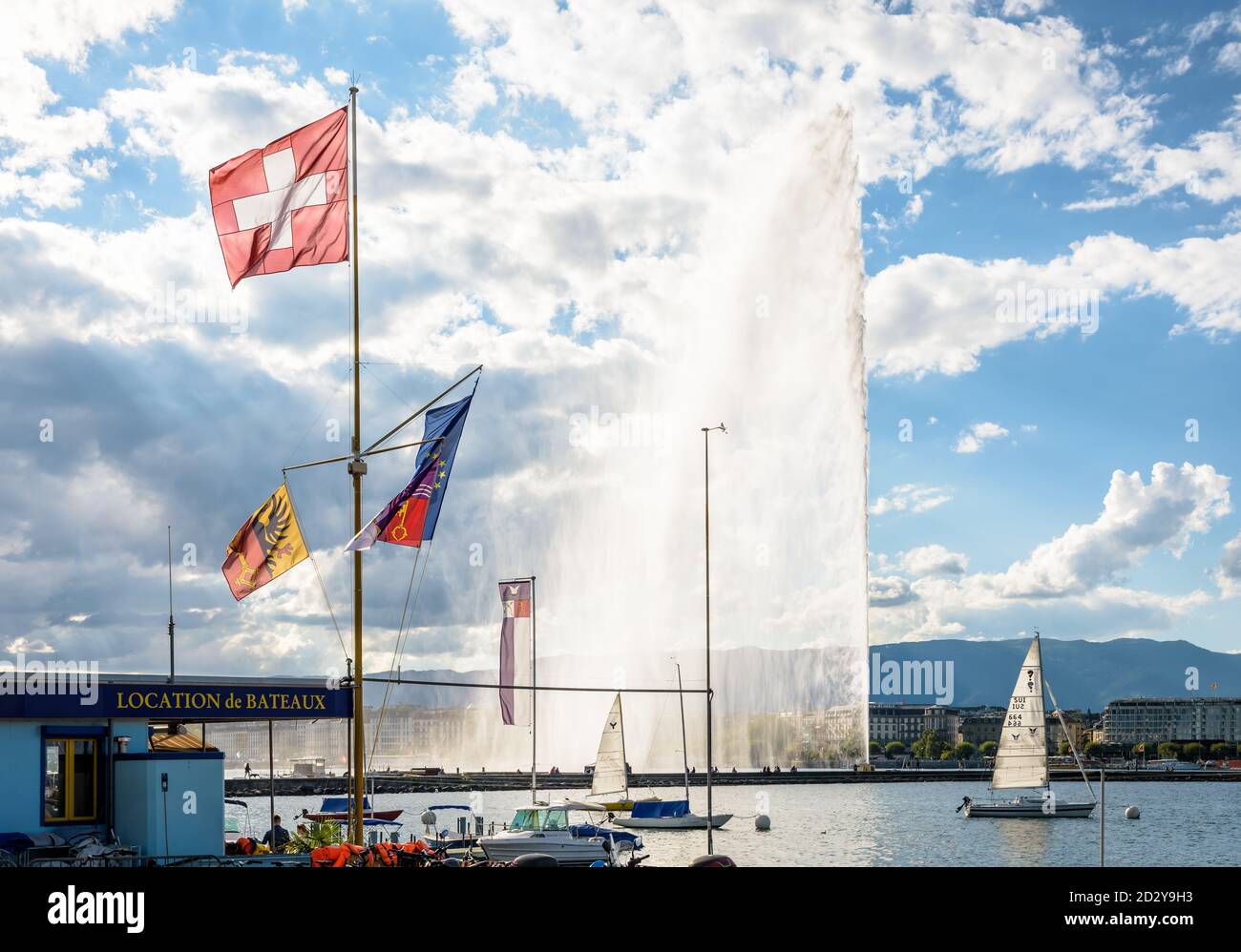 Die Schweizer Flagge fliegt an einem stürmischen Tag am Ufer des Genfersees in Genf, Schweiz, mit dem Jet d'Eau Wasserstrahlbrunnen in der Ferne Stockfoto