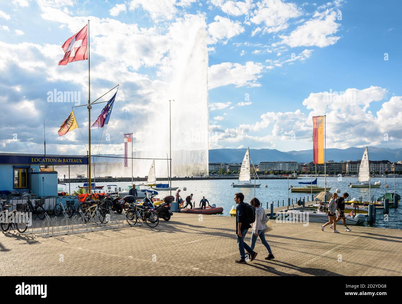 Menschen, die am Genfersee in Genf, Schweiz, spazieren, mit der Schweizer Flagge im Vordergrund und dem Jet d'Eau Wasserstrahlbrunnen in der Ferne Stockfoto